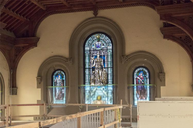 Behind scaffolding, an end wall contains three arched windows each containing a stained glass window.