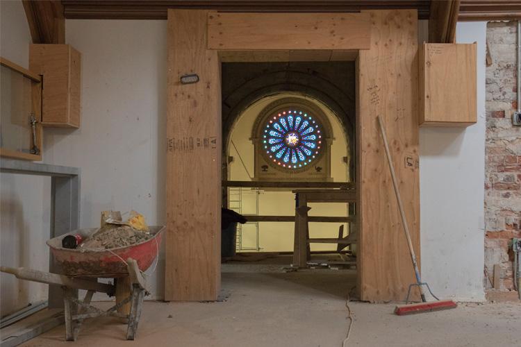 A wheelbarrow filled with rubble sits next to a door shored up with plywood. Through the door, a stained glass window glows.