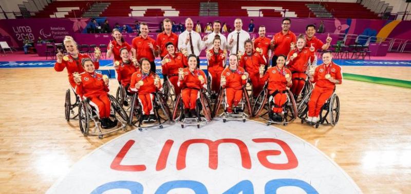 Athletes sitting in wheelchairs on a basketball court smile and hold up two medals each.