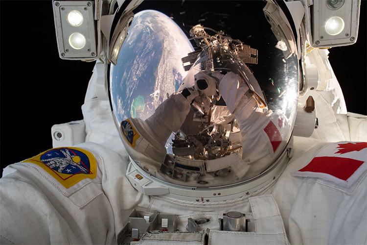 A photo of David Saint-Jacques in a space suit shows the earth and his camera reflected in his helmet.