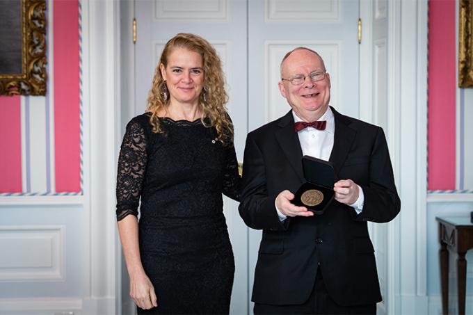 Andrew James Paterson smiles and holds up his medal while standing next to a smiling Julie Payette.