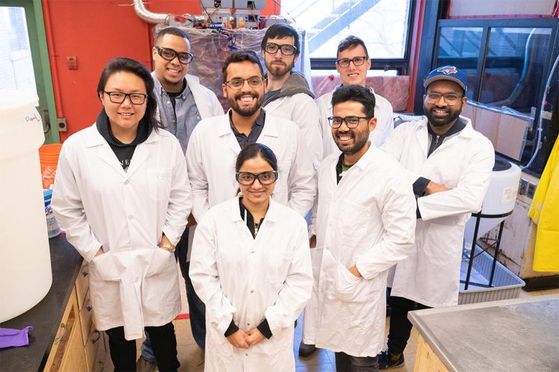 A diverse team of people wearing lab coats and goggles smile up from the floor of a lab filled with large machines.