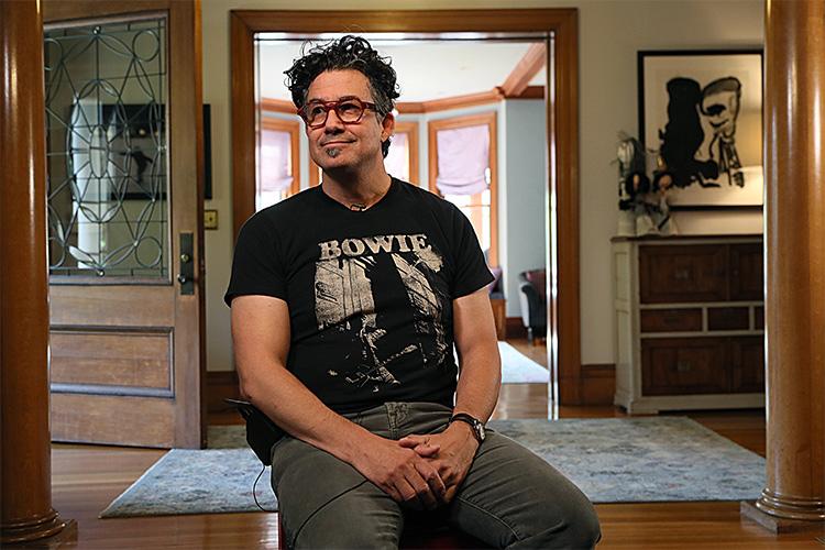 Derrick Rossi smiles as he sits in the hallway of a nice house, wearing a Bowie T-shirt.