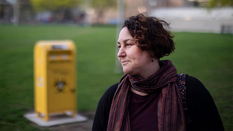 Gillian Kolla looks serious as she stands in a park with a needle disposal bin behind her.