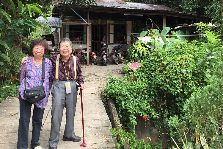 Eng and C.C. Liew smile as they stand in the lush garden of a small house with a patched metal roof.