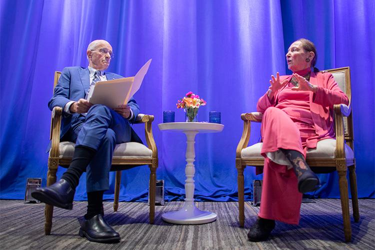 Rosalie Abella gestures as she sits in a chair on stage facing Frank Iacobucci, who is listening intently.