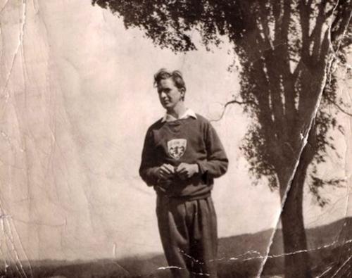 In a cracked old photo from perhaps the 1930s, a young Lou Siminovitch stands under a tree and wears a sweater with a crest.