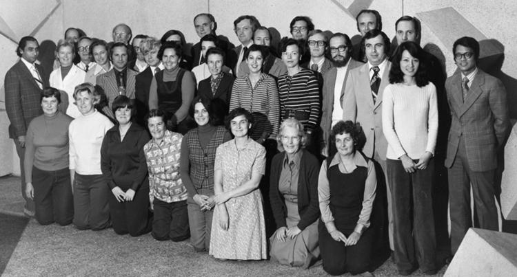 In a photograph from 1976, a group of smiling people pose in rows in a room decorated in funky abstract shapes.