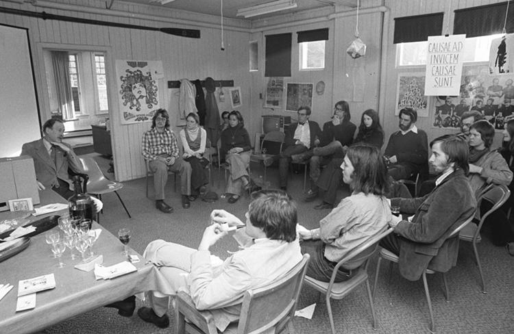 In an image from the 1970s, a group of students sit in a semicircle around Marshall McLuhan. Wineglasses sit on the table.