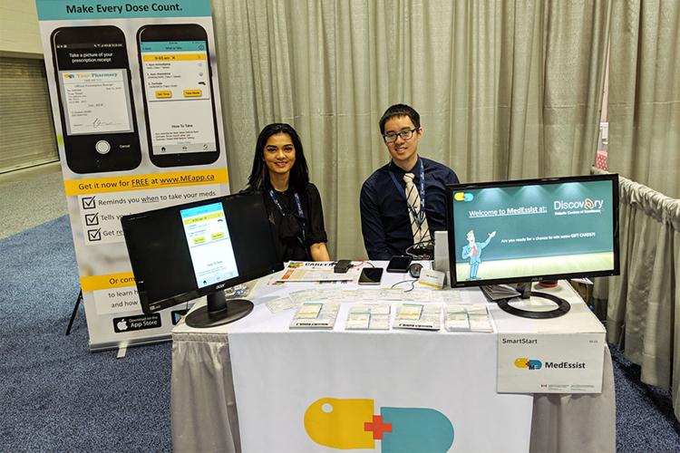 Joella Almeida and Michael Do smiles as they sit at a conference booth. Displays and leaflets illustrate their app MedEssist.