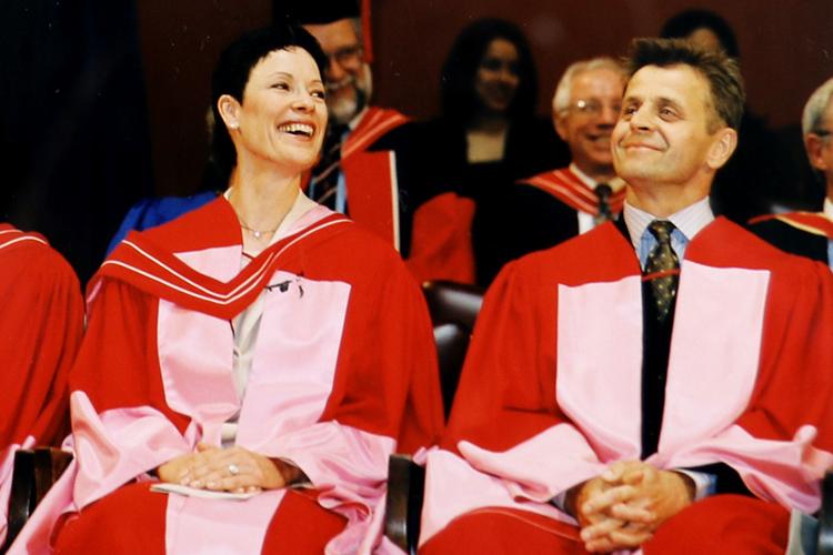 Karen Kain and Mikhail Baryshnikov share a laugh together as they sit on stage wearing U of T academic robes.