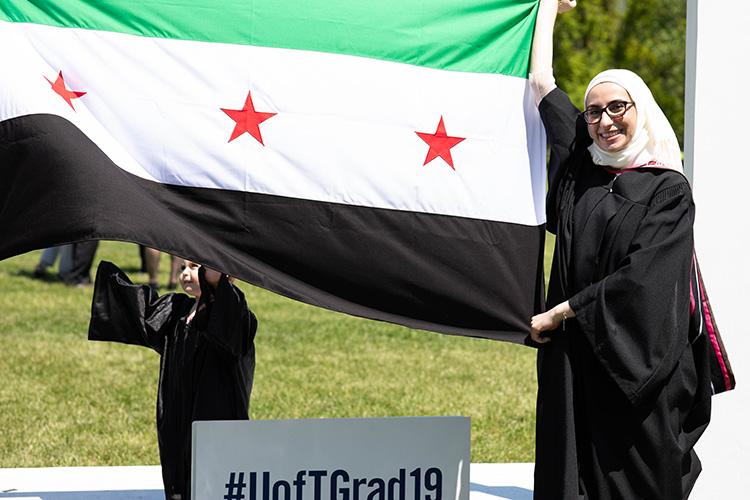 Noura Al-Jizawi and her small daughter smile and hold up a large Syrian independence flag. Both are wearing academic robes.