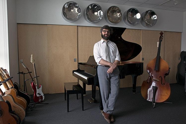 Richard Marsella smiles as he leans casually on a grand piano in a room filled with guitars and a double bass.
