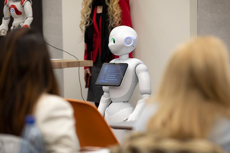 A human-shaped, child-sized robot with a text panel on its chest stands in front of an audience