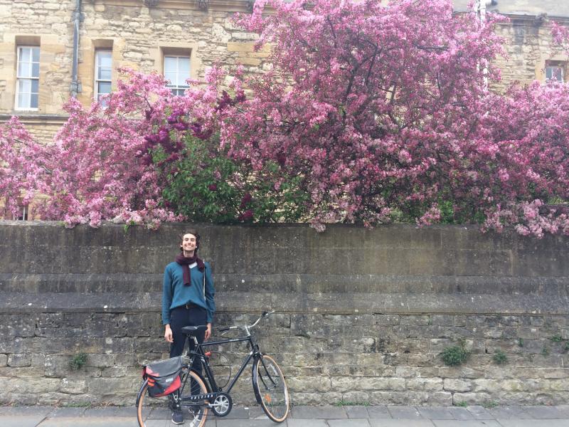 Phil Schwarz smiles as he stands with his bicycle by a stone wall. The wall is topped with a cloud of pink flowers.