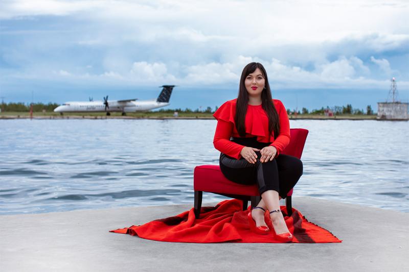 Lesley Hampton smiles as she poses in a dramatic ruffled shirt, on a dock across from a lakeside airport.