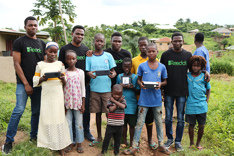 Four men wearing Reedi T-shirts pose with a group of children holding Reedi devices in a village among trees.