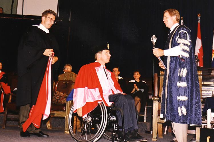 Rick Hansen, wearing academic robes, wheels his chair up to U of T's President on stage at Convocation Hall.