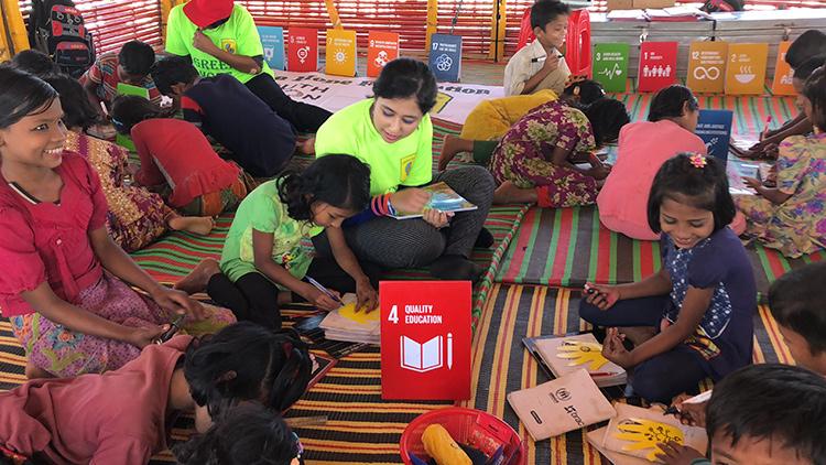 Kehkashan Basu sits among children on mats in a tent. The kids are tracing and decorating hand cut-outs.