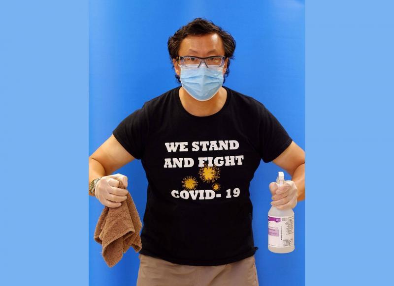 Stephen Lew wears a mask and a T-shirt reading We Stand and Fight COVID-19. He holds cleaning supplies in his gloved hands.