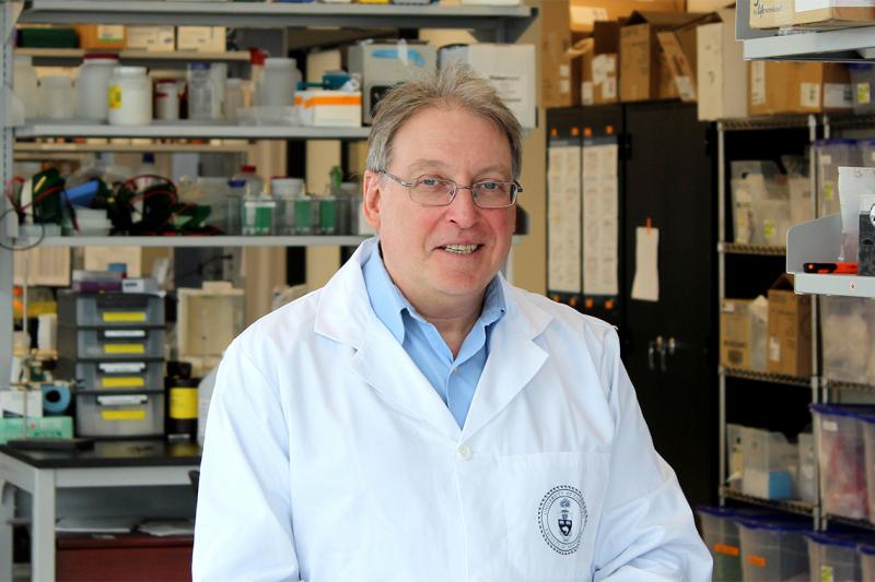 Paul Santerre smiles as he stands among shelves of lab equipment, wearing a U of T lab coat.