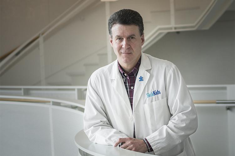 Stephen Scherer has a slight smile as he wears a SickKids lab coat and leans on a railing indoors.