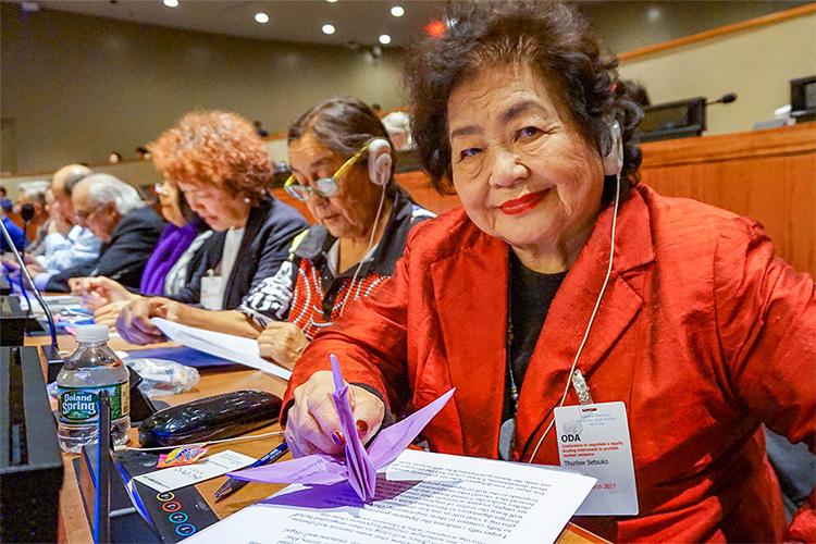 Setsuko Thurlow smiles and holds up a paper crane as she sits among a row of delegates in a conference amphitheatre.