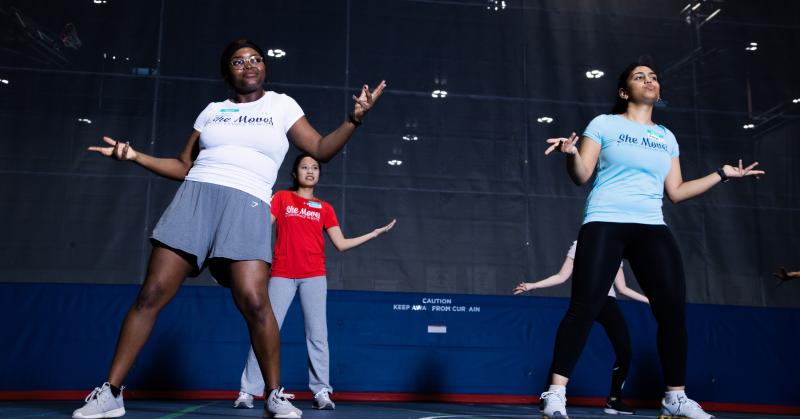 4 women wearing workout gear try a Bollywood dance move with feet apart and hands gesturing.