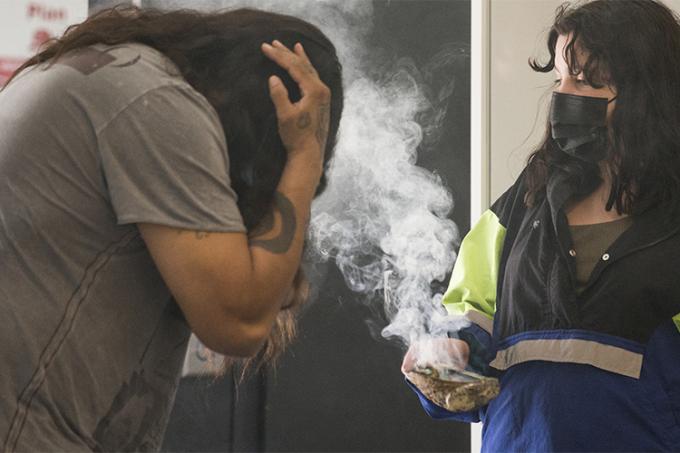 A woman holds a shell with a plume of smudging smoke rising from it. A man leans forward to hold his head in the smoke.
