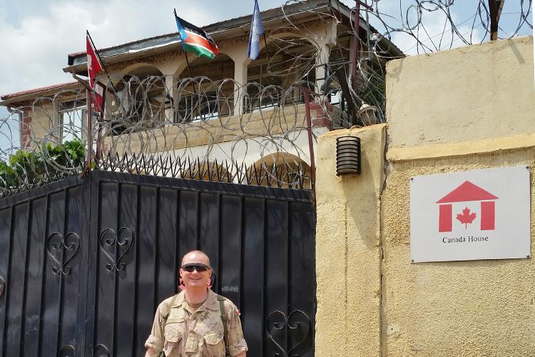 Wearing desert fatigues, Gary Johnston stands by a mud-brick wall topped with razor wire and a sign that reads Canada House.