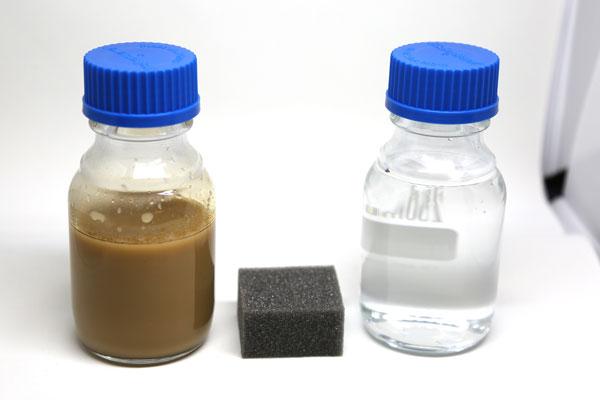 A small square sponge sits between a beaker of filthy water and a beaker of clean water.