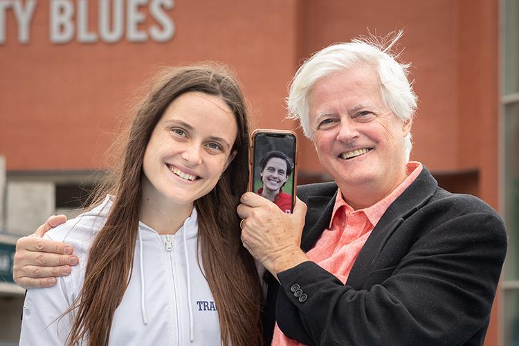 Lucia and Jamie Stafford smile and hold up a phone showing Gabriela DeBues-Stafford on the video screen.