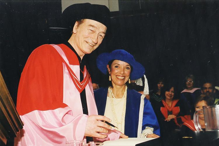 Stompin Tom Connors smiles as he signs a book in Convocation Hall. He is wearing a cowboy hat and academic robes.
