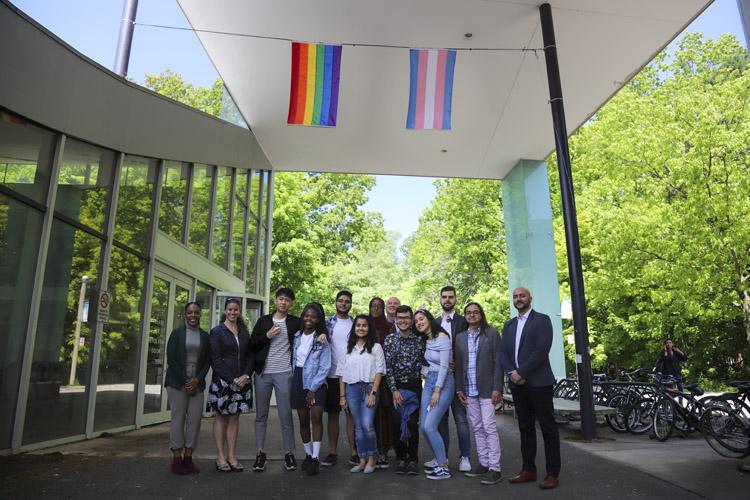 A diverse group of students, staff and faculty members stand under the Pride and Trans flags at the entrance to the UTM Student Centre.