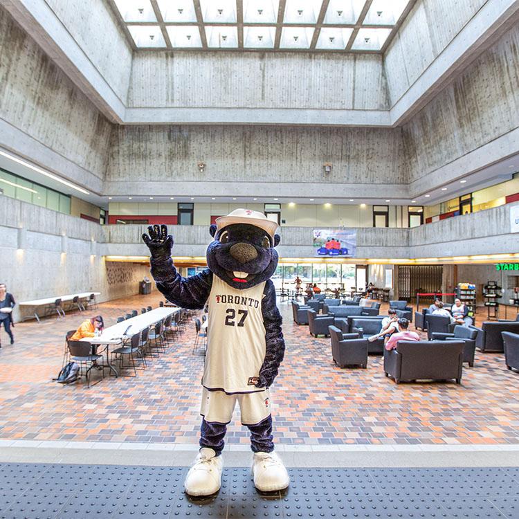 The True Blue mascot waves while standing under the skylight and concrete balconies of UTSC's Meeting Place.