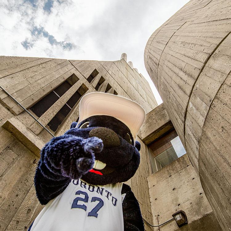 The True Blue mascot points at the camera in front of a background of a tall concrete tower topped with three smokestacks.