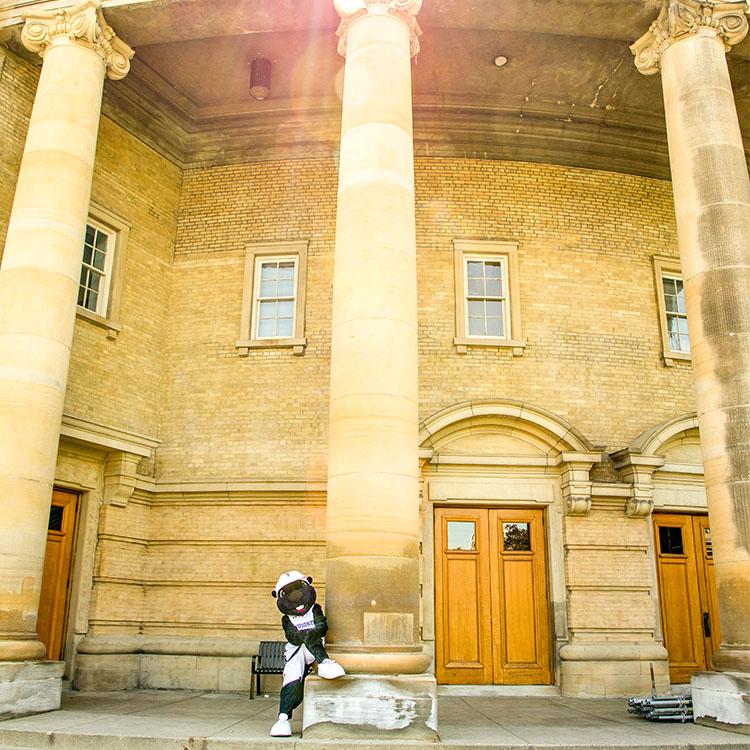 The True Blue mascot props one foot on the base of a tall pillar outside Convocation Hall.