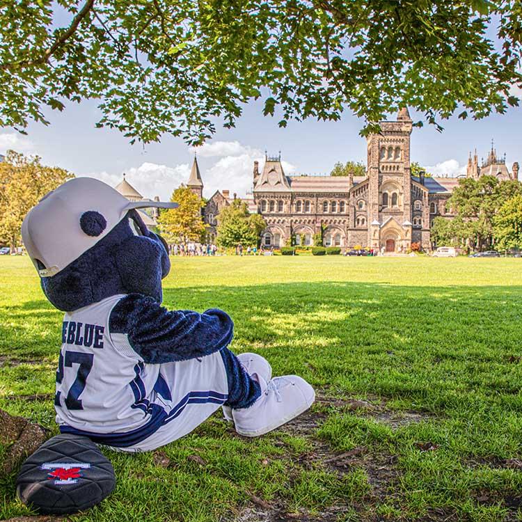 The True Blue mascot sits on the grass under a tree, arms around knees, and looks over Front Campus at University College.