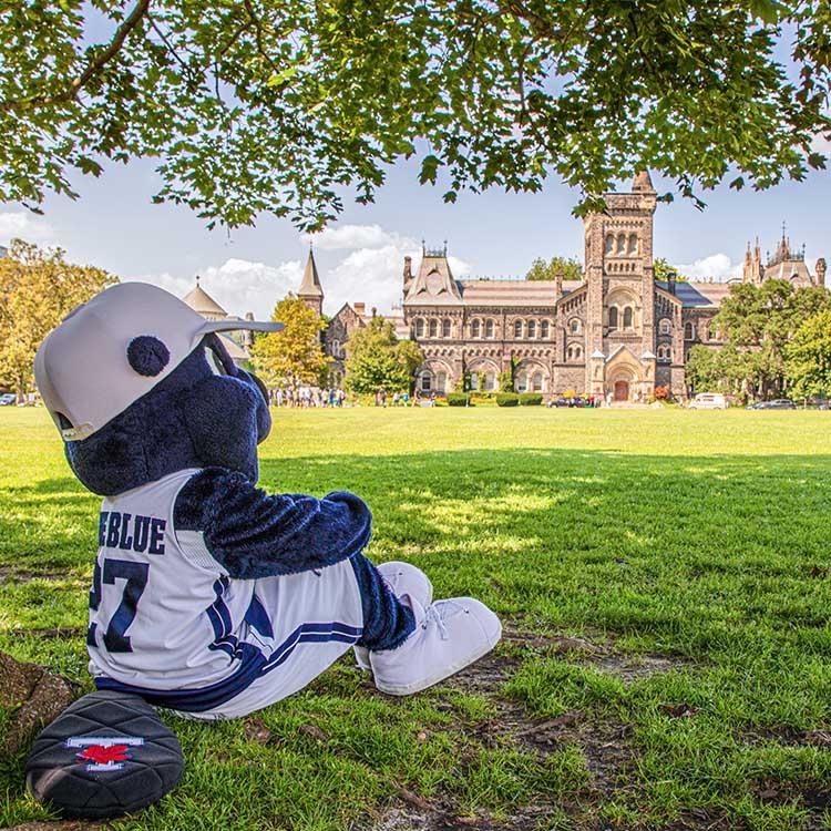 The True Blue mascot sits on the grass under a tree, arms around knees, and looks over Front Campus at University College.