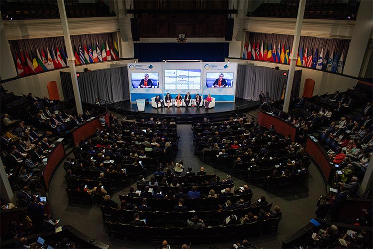Viewed from above, the seats of Convocation Hall are filled. Onstage, six panellists sit in armchairs before an array of flags.
