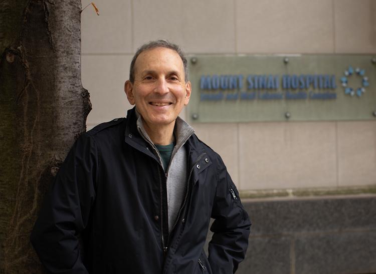 Daniel Druckers smiles happily, leaning on a tree by a sign that reads Mount Sinai Hospital.