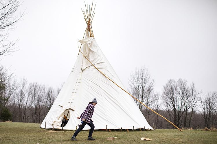 A young woman chops wood outside a large canvas tipi. Behind, a wood of winter trees stretches away.