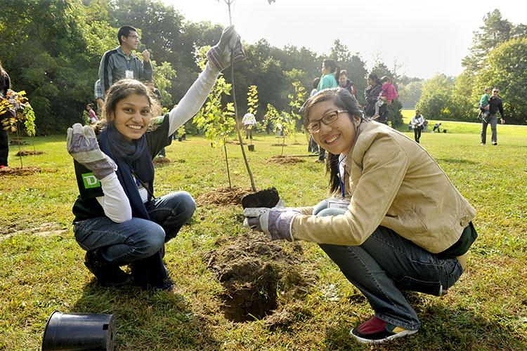 Two young women smile as they squat beside a hole in a lawn and hold up a tree seedling.