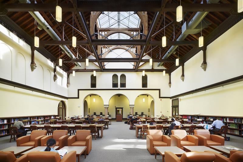 An enormous hall features a large central skylight. Below, study tables and comfy chairs are placed between bookshelves.