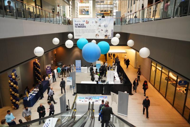 Escalator with people leading down to the True Blue Expo event. Blue and white balloons above the escalators and large signage reading "The Place to Innovate."