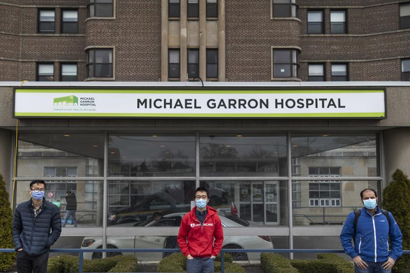 Joseph Choi, Albert Tai and Umar Azhar wear masks and stand two metres apart in front of a sign reading: Michael Garron Hospital