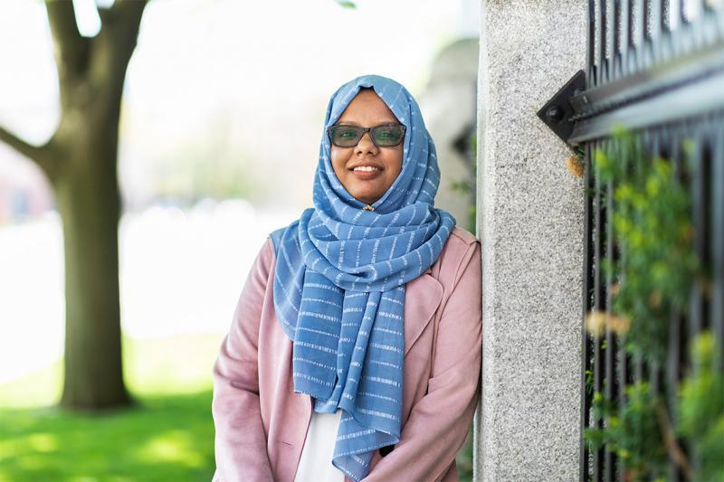 Mariam Abeid smiles as she leans on an iron fence near a tree outdoors.