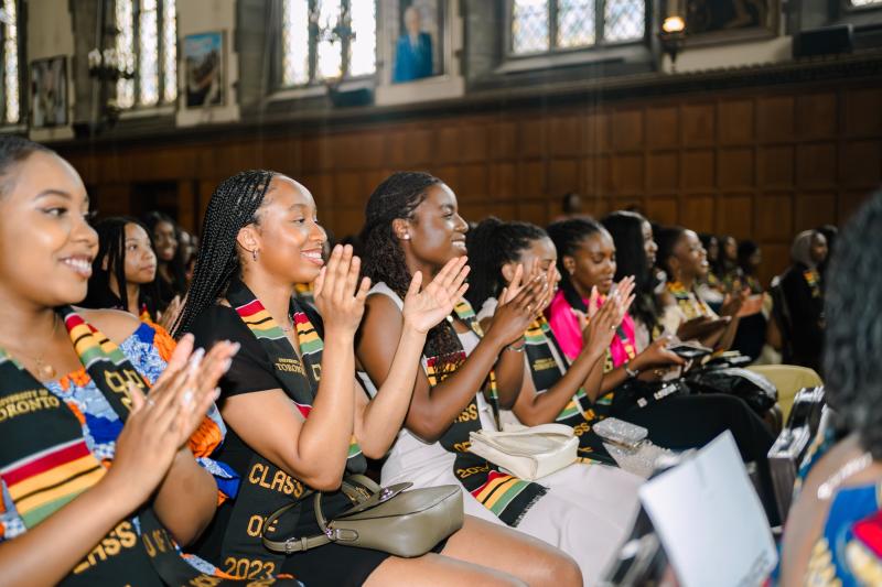 A group of Black Graduates sits in Hart House applauding fellow graduates.
