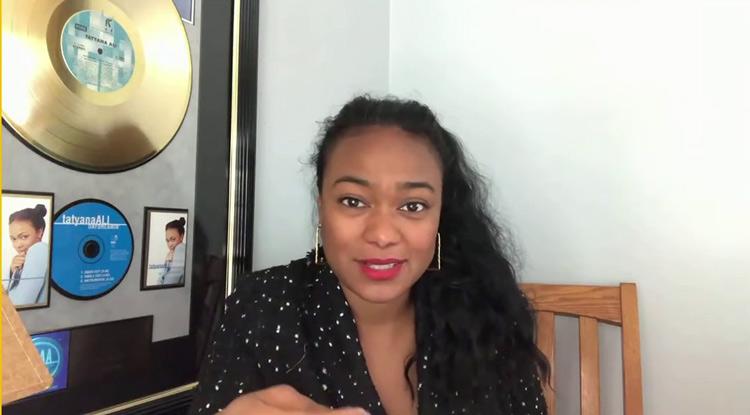 Tatyana Ali smiles as she talks on a video call, with music awards displayed behind her.