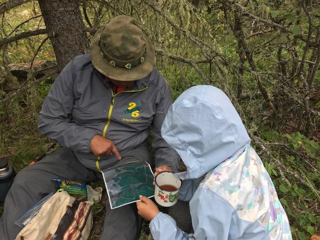 An older man and a young woman sit together on the ground under a small tree and lean over a map of woods and rivers.